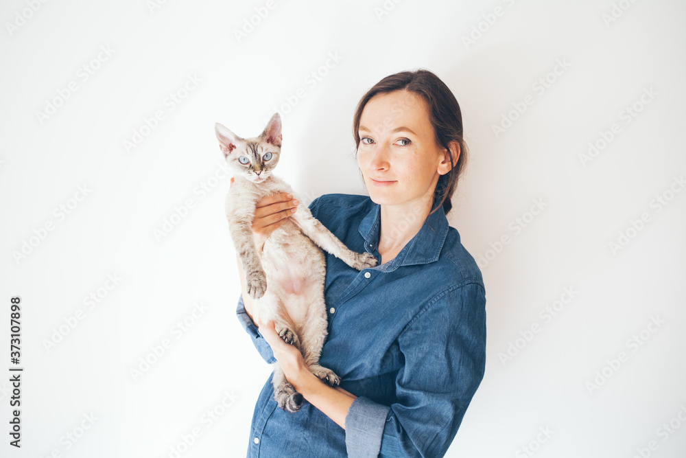 Obraz premium Lifestyle photo of a casual dressed female in blue jeans shirt holding and cuddling cute and funny Devon Rex cat. Happy kitty in girls arms. Breed with special hypoallergenic, no low shedding fur