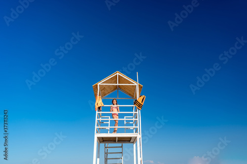 teenage girl stands on lifeguard tower on beach against cloudless sky.
