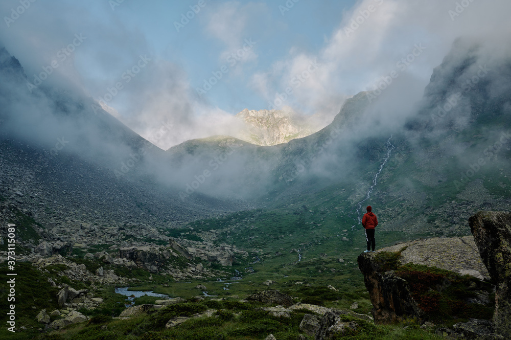 A young man in the red jacket stands on the rock against the mountain range among the clouds. An extreme hiker in the wild nature. Domestic travel trekking lifestyle. Local tourism