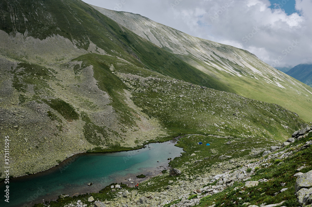 A mountain valley landscape with green hills, turquoise lake and white clouds. Two little blue tents among majestic wild nature. Domestic travel trekking. Local tourism