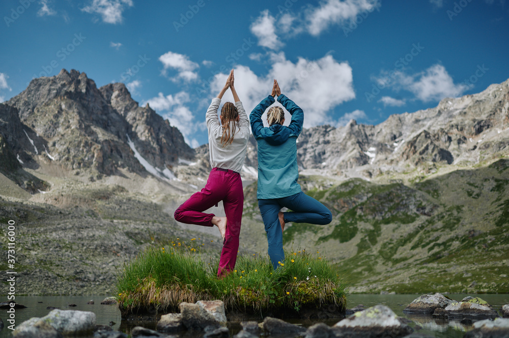 Two women stay in one leg yoga position against the turquoise lake in the mountains. Extreme hiker girl rest in the wild nature. Trekking lifestyle. Domestic local hike, tourism, travel