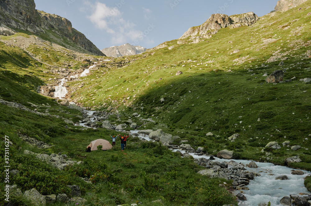 Three hikers friends are staying by the tourist tend and river among green mountains. Extreme tourists in wild nature landscape. Domestic travel trekking lifestyle. Local tourism Three hikers friends are staying by the tourist tend and river among green mountains. Extreme tourists in wild nature landscape. Domestic travel trekking lifestyle. Local tourism