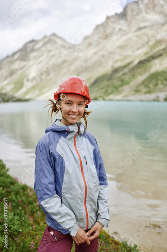 Young hiker woman portrait. Adult girl in helmet is standing by the mountain lake shore and smiling at camera. Domestic travel and trekking. Local tourism. Healthy lifestyle © kravtzov