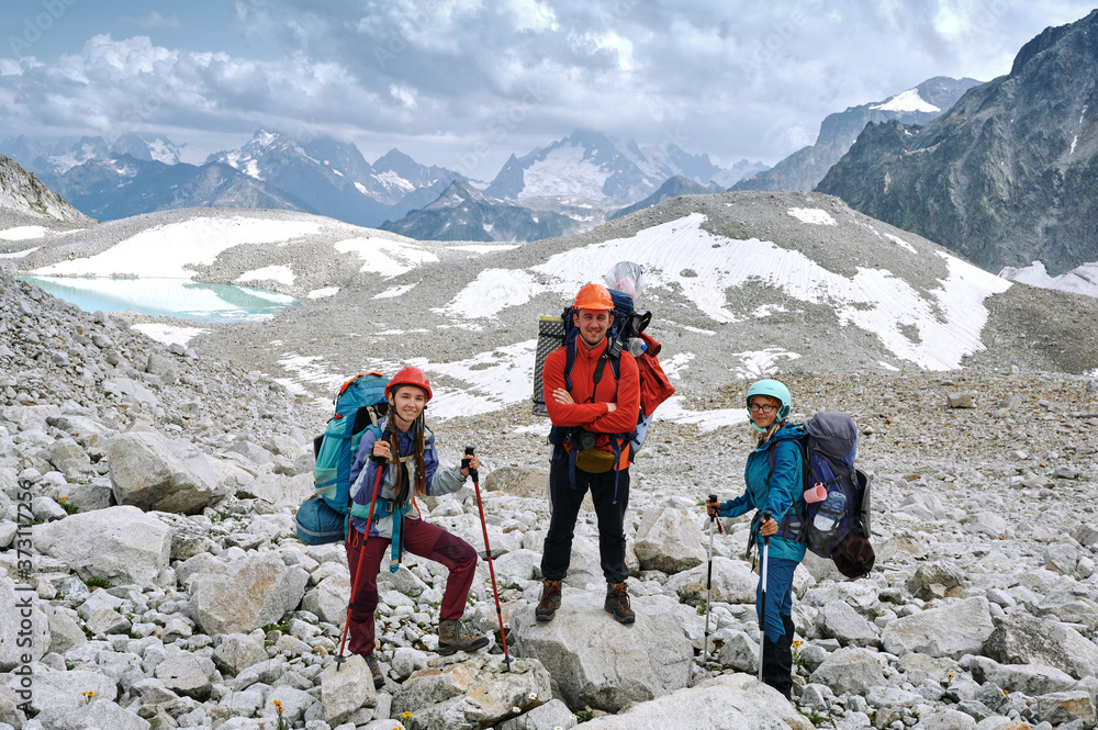 Three hikers with backpacks and helmets are staying among the rocky mountain valley and looking at camera. Extreme backpacker tourist in wild nature. Domestic travel and trekking. Local tourism.