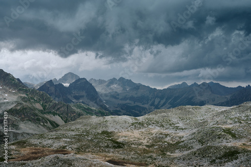 Gloomy mountain landscape with thunder cloudy sky, rocky ranges and peaks with glaciers and snow fields. Wild nature valley