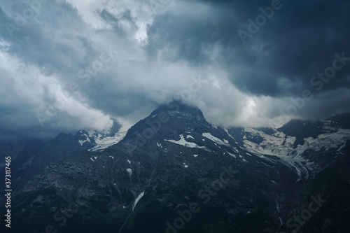 Gloomy mountain top landscape with thunder cloudy sky, rocky ranges and peaks with glaciers and snow fields. Rainy day in wild nature. Belalakaya, Dombay, Caucasus