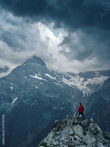 Alone hiker man in red jacket against the gloomy mountain top landscape with thunder cloudy sky, rocky ranges and peaks with glaciers and snow fields. Domestic travel and trekking. Local tourism.