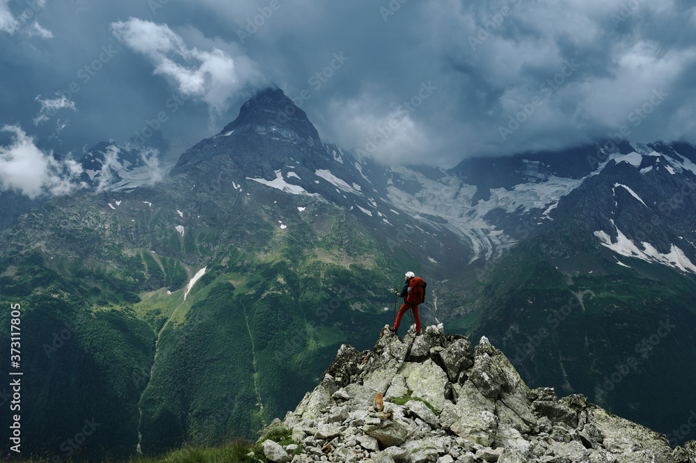 Obraz premium Alone hiker man with backpack and helmet against the gloomy mountain peak landscape with thunder cloudy sky, rocky ranges and peaks with glaciers. Extreme domestic travel and trekking. Local tourism.