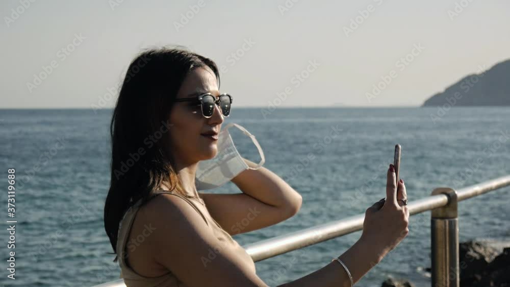 Girl takes off protective face mask making selfie with smartphone on sea pier. The new normal concept. Girl takes off protective face mask making selfie with smartphone on sea pier. The new normal concept.