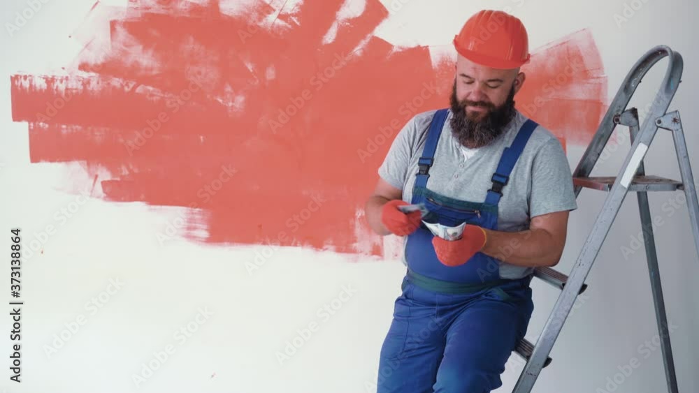 A bearded man is wearing a jumpsuit uniform and a protective helmet. The builder is sitting on the stairs and counting money. a stack of 100 bills A bearded man is wearing a jumpsuit uniform and a protective helmet. The builder is sitting on the stairs and counting money. a stack of 100 bills