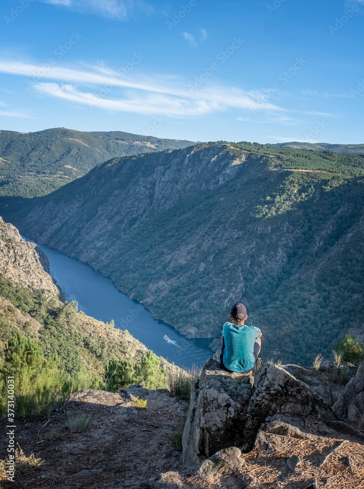boy contemplating a ship sailing through a canyon sitting on top of a cliff in the canyon of the river Sil in the Ribeira Sacra of Lugo, Spain boy contemplating a ship sailing through a canyon sitting on top of a cliff in the canyon of the river Sil in the Ribeira Sacra of Lugo, Spain