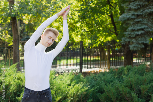 A young blond guy with closed eyes, dressed in a white sweater and black jeans, holds his hands up, clasping them against the backdrop of a city park. Sports concept.