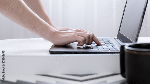 Trabajando en el ordenador portátil desde la comodidad del hogar. Primer plano de manos caucásicas de hombre escribiendo sobr eun teclado negro de una laptop en una habitación blanca e iluminada