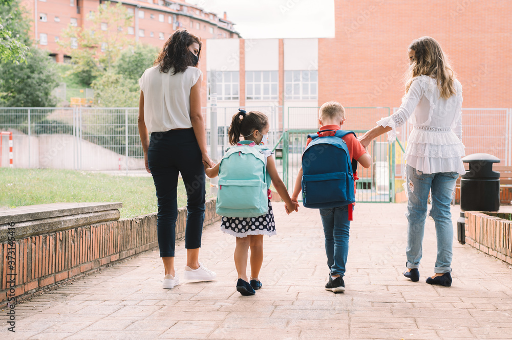 Two women and two children holding hands from back go to school for first time after coronavirus pandemic. Mothers accompanies the students along way. Two women and two children holding hands from back go to school for first time after coronavirus pandemic. Mothers accompanies the students along way.