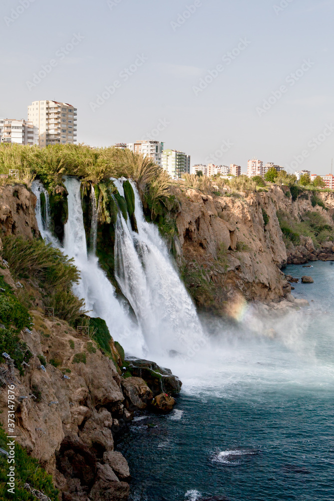 A waterfall erupts into the sea from a great height on a sunny day near the city A waterfall erupts into the sea from a great height on a sunny day near the city