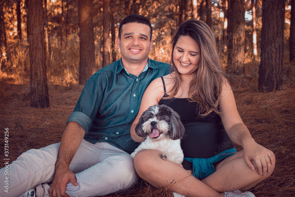 Pregnant woman sitting on the floor of a pine field, accompanied by a man and a dog. Reddish autumn photo. Family with dog and pregnant woman