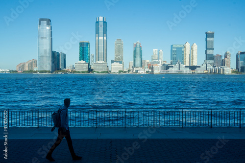 New York, NY / USA - 8/20/20: an image of a commuter walking on the Battery Park City Esplanade with the Hudson River and the Jersey City skyline behind him © Brian