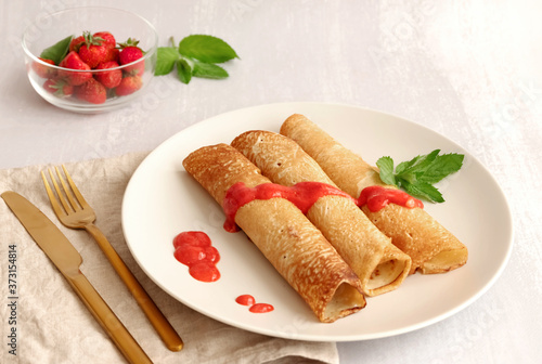 Three delicious crepes on a plate covered with strawberry sauce, light grey background 