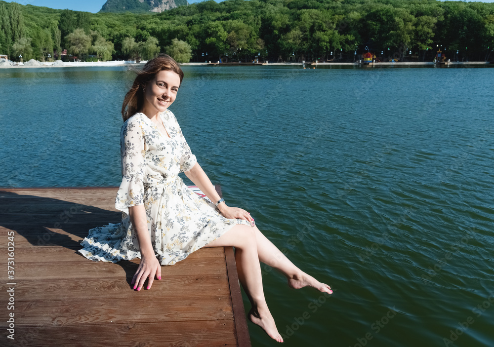 A beautiful and sexy woman, with long hair in a summer dress, spends time on the shore of the lake, surrounded by forests and mountains. Rest and relaxation concept