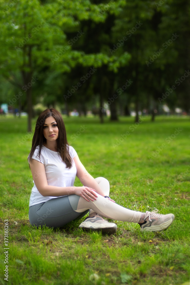 Pretty woman in sports wear sitting on city park on green lawn