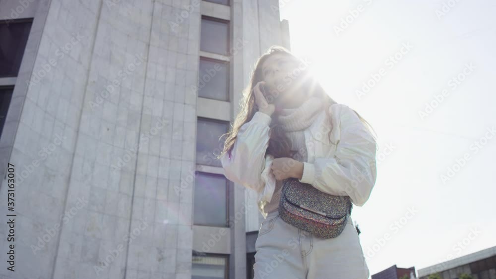 A dark-haired young woman standing near the building and having a conversation with her friend on the phone. A slow motion bottom view close up A dark-haired young woman standing near the building and having a conversation with her friend on the phone. A slow motion bottom view close up