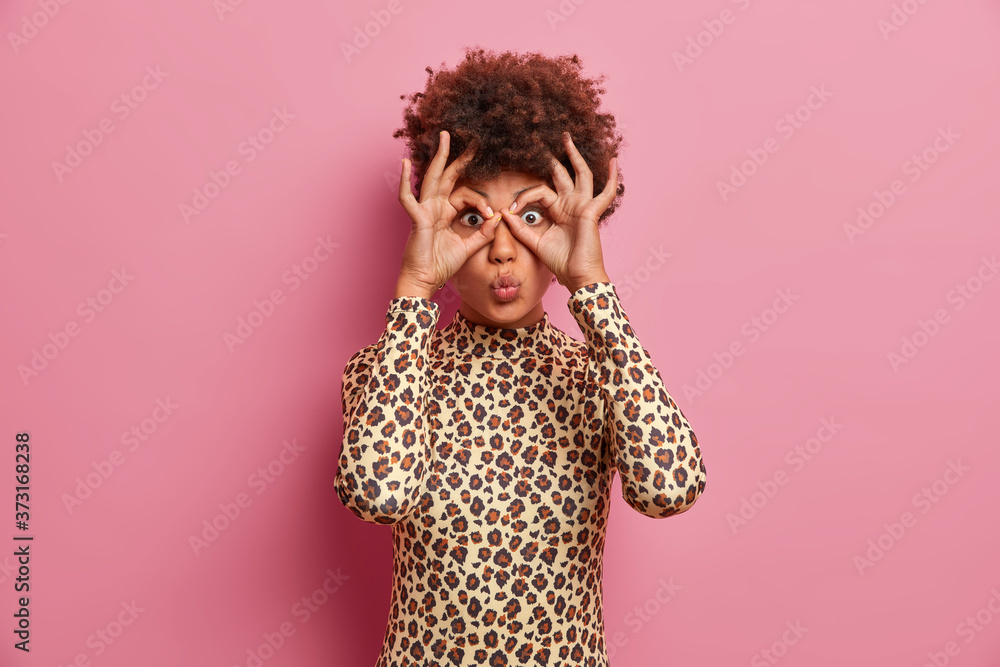 Surprised dark skinned woman in leopard jumper makes goggles with fingers, folds lips and foolishes around, has fun, makes glasses or okey gesture, isolated over rosy background, enjoys free time Surprised dark skinned woman in leopard jumper makes goggles with fingers, folds lips and foolishes around, has fun, makes glasses or okey gesture, isolated over rosy background, enjoys free time
