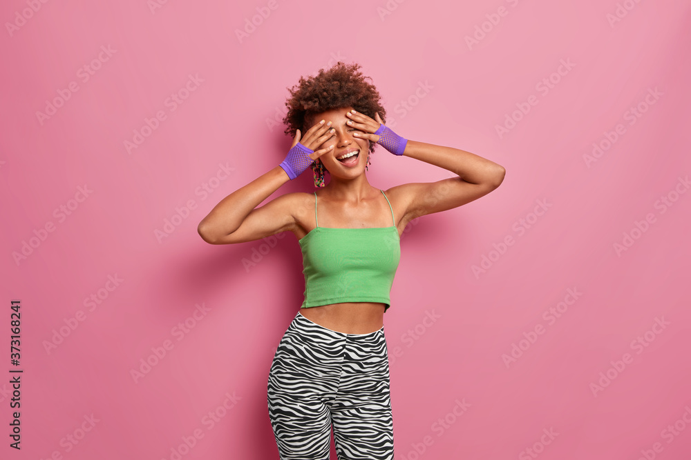 Horizontal shot of positive carefree slim woman covers face with hands, smiles broadly, keeps eyes shut, wears sport clothing, goes in for sport regularly, being energetic, poses over pink background Horizontal shot of positive carefree slim woman covers face with hands, smiles broadly, keeps eyes shut, wears sport clothing, goes in for sport regularly, being energetic, poses over pink background