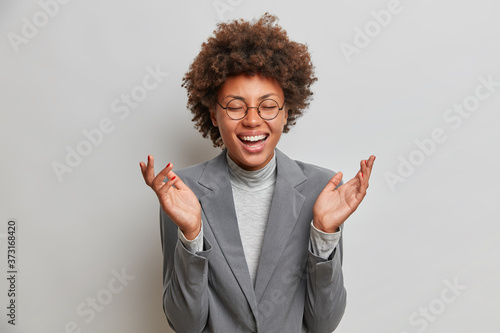 Carefree overjoyed woman office worker laughs joyfully, amused by funny colleagues, closes eyes from happiness, wears formal clothes, hears funny story, has upbeat mood, isolated on grey wall