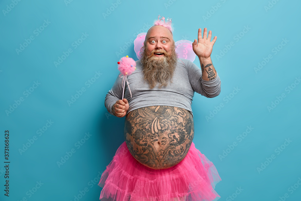 Fototapeta premium Horizontal positive man animator on childrens party, dressed in fairy costume, waves palm in hello gesture, has good mood during holiday, being kind magician, has big tattooed belly, blue background