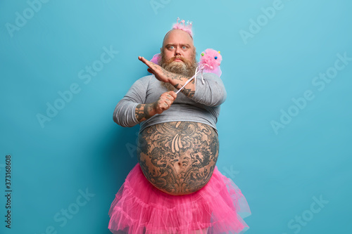 Photo of serious bearded man with big belly, makes refusal gesture, wears fairy costume, refuses to go on stage in funny outfit, crosses hands, poses against blue background. Party time concept