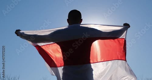 A masked man holds an unfurled Belarus flag and slightly raises it up in the backlight of the sun. photo