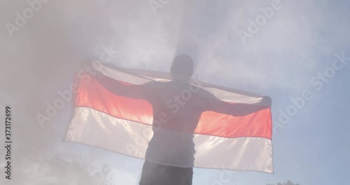 Belarus in a mask and sunglasses holds the flag of Belarus stretched in his hands in the backlight of the sun against a background of smoke. photo