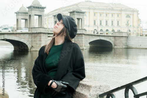 Stylish long-haired Girl in a green dress, black coat and a cap smiles on the Fontanka river embankment © Дэн Едрышов