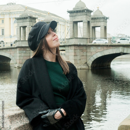 Stylish long-haired Girl in a green dress, black coat and a cap smiles on the Fontanka river embankment © Дэн Едрышов