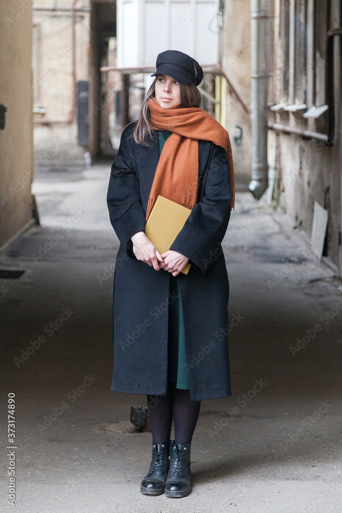 Long-haired girl student in a black coat with a vintage cap and a red scarf, posing with a book in a shabby quiet courtyard