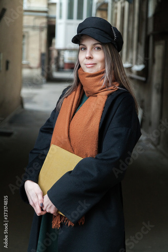 Long-haired girl student in a black coat with a vintage cap and a red scarf, posing with a book in a shabby quiet courtyard © Дэн Едрышов