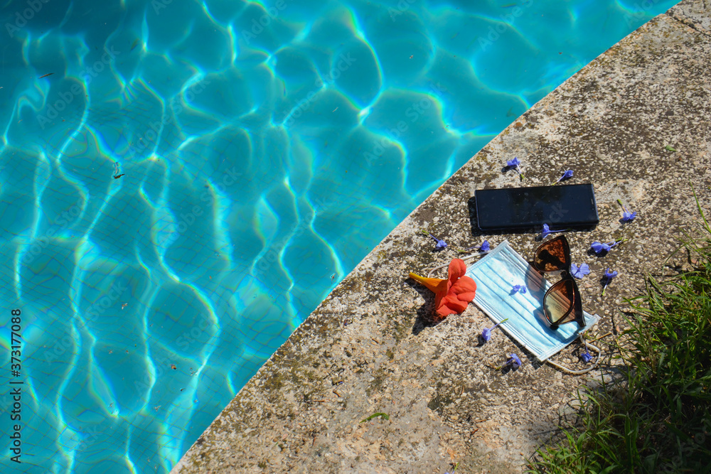 Blue face mask for protection against the Corona Virus (COVID-19), sunglasses and cell phone laying beside a swimming pool during coronavirus worldwide pandemic holidays