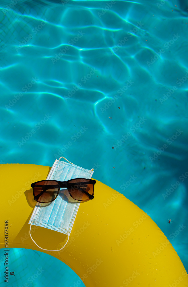 Blue face mask for protection against the Corona Virus (COVID-19), sunglasses laying on a pool mat beside a swimming pool during coronavirus worldwide pandemic holidays