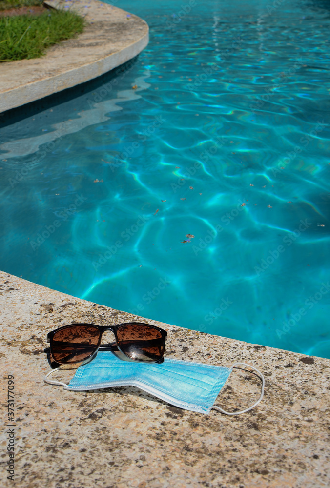 Blue face mask for protection against the Corona Virus (COVID-19), sunglasses laying beside a swimming pool during coronavirus worldwide pandemic holidays Blue face mask for protection against the Corona Virus (COVID-19), sunglasses laying beside a swimming pool during coronavirus worldwide pandemic holidays