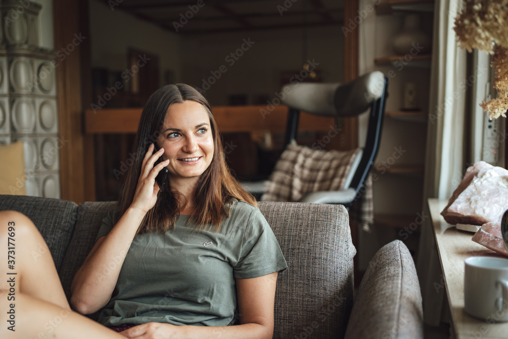 Obraz premium Young Caucasian woman looking away while talking on mobile phone, sitting on sofa at home