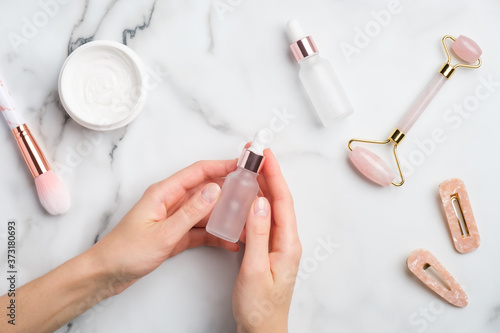Female hand holding serum bottle over marble table with moisturizer cream, makeup brush, face massage roller. Flat lay, top view. Natural organic essential oil, skin care beauty product.