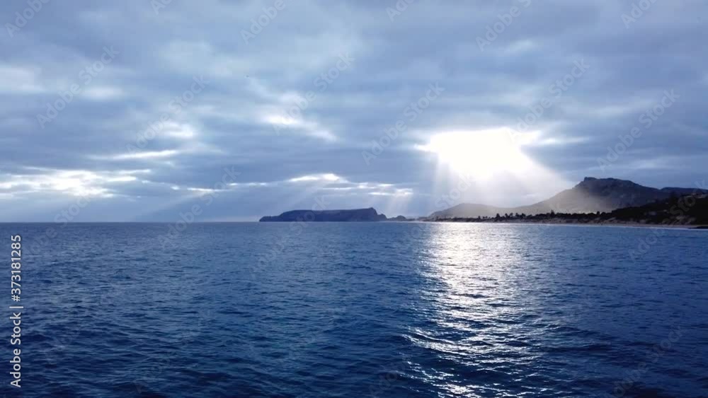 Hyperlapse: spectacular sun rays and beams shining through opening in blanket of grey stormy clouds in sky above blue ocean sea waters by Porto Santo island beach and mountain range, pan right Hyperlapse: spectacular sun rays and beams shining through opening in blanket of grey stormy clouds in sky above blue ocean sea waters by Porto Santo island beach and mountain range, pan right