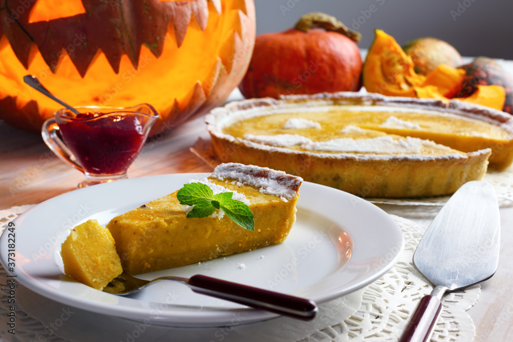 Piece of homemade halloween pumpkin cake with leaf of mint on white plate. Jack-o-lantern, fork and cowberry jam around Piece of homemade halloween pumpkin cake with leaf of mint on white plate. Jack-o-lantern, fork and cowberry jam around