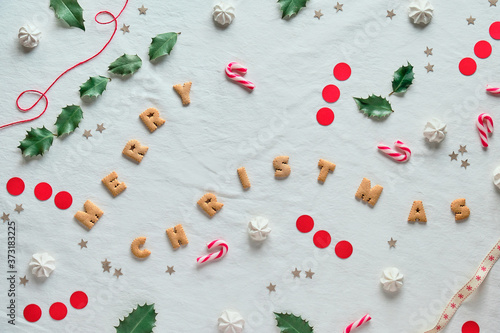 Eco-friendly Xmas decor with natural holly leaves. Flat lay on white textile. Merry Christmas text made of cookies. Xmas decor silhouettes cut out from brown and red paper and sweet marshmallows.