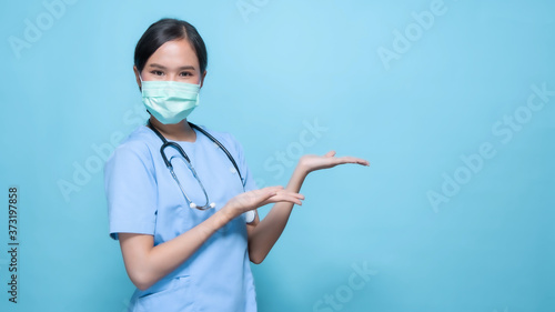 Portrait of young Asian beautiful nurse in blue uniform and wearing a surgical mask for protection from covid19 or disease with big smiled inside pointing and present on copy space blue background