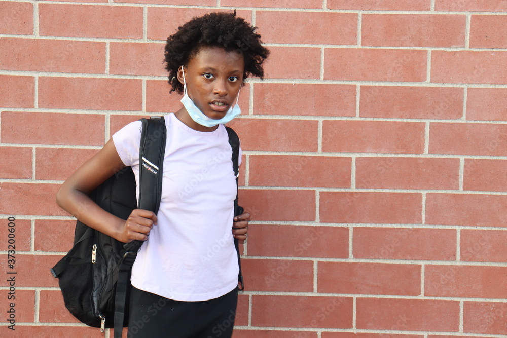 Black girl wearing backpack and surgical face mask pulled down standing outdoors red brick background Black girl wearing backpack and surgical face mask pulled down standing outdoors red brick background