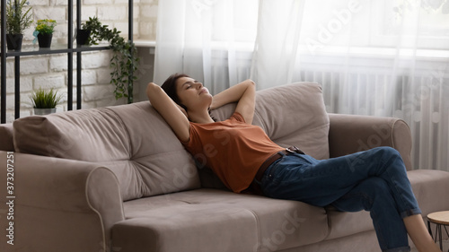 Young Caucasian woman sit relax on comfortable sofa in living room sleep or take nap at home, millennial girl rest on couch relieve negative emotions, breathe fresh air, stress free, peace concept