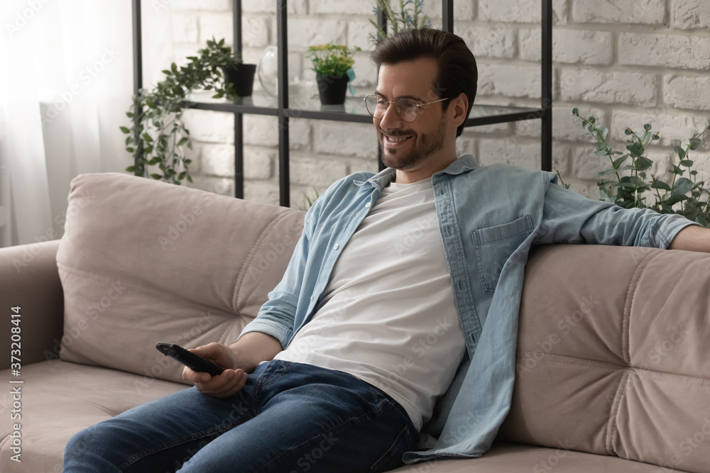 Smiling young man sit rest on couch in living room hold remote controller watching TV at home, happy millennial Caucasian guy relax on sofa enjoy smart television program, weekend concept Smiling young man sit rest on couch in living room hold remote controller watching TV at home, happy millennial Caucasian guy relax on sofa enjoy smart television program, weekend concept