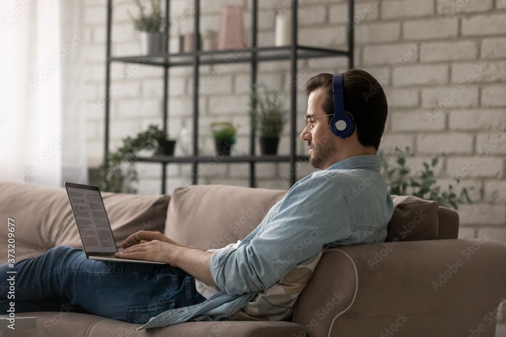 Young Caucasian man in headphones sit on couch in living room work online on laptop, male in earphones busy studying preparing report project on computer at home office, use wireless internet Young Caucasian man in headphones sit on couch in living room work online on laptop, male in earphones busy studying preparing report project on computer at home office, use wireless internet