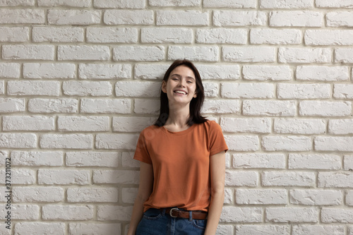Portrait of positive millennial Caucasian girl teenager isolated on white brick wall look at camera smiling, happy young woman tenant or renter feel optimistic overjoyed, show white healthy teeth